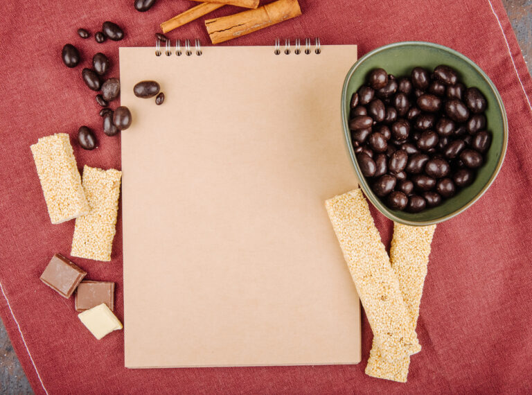top view of a sketchbook and glazed chocolate nut candies in a bowl with pieces of sesame kozinaki on dark pink tablecloth
